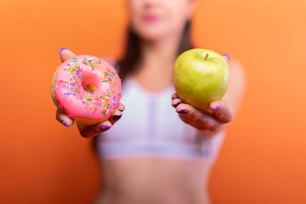 A woman holds a green apple and a pink donut with sprinkles against an orange background, emphasizing choice